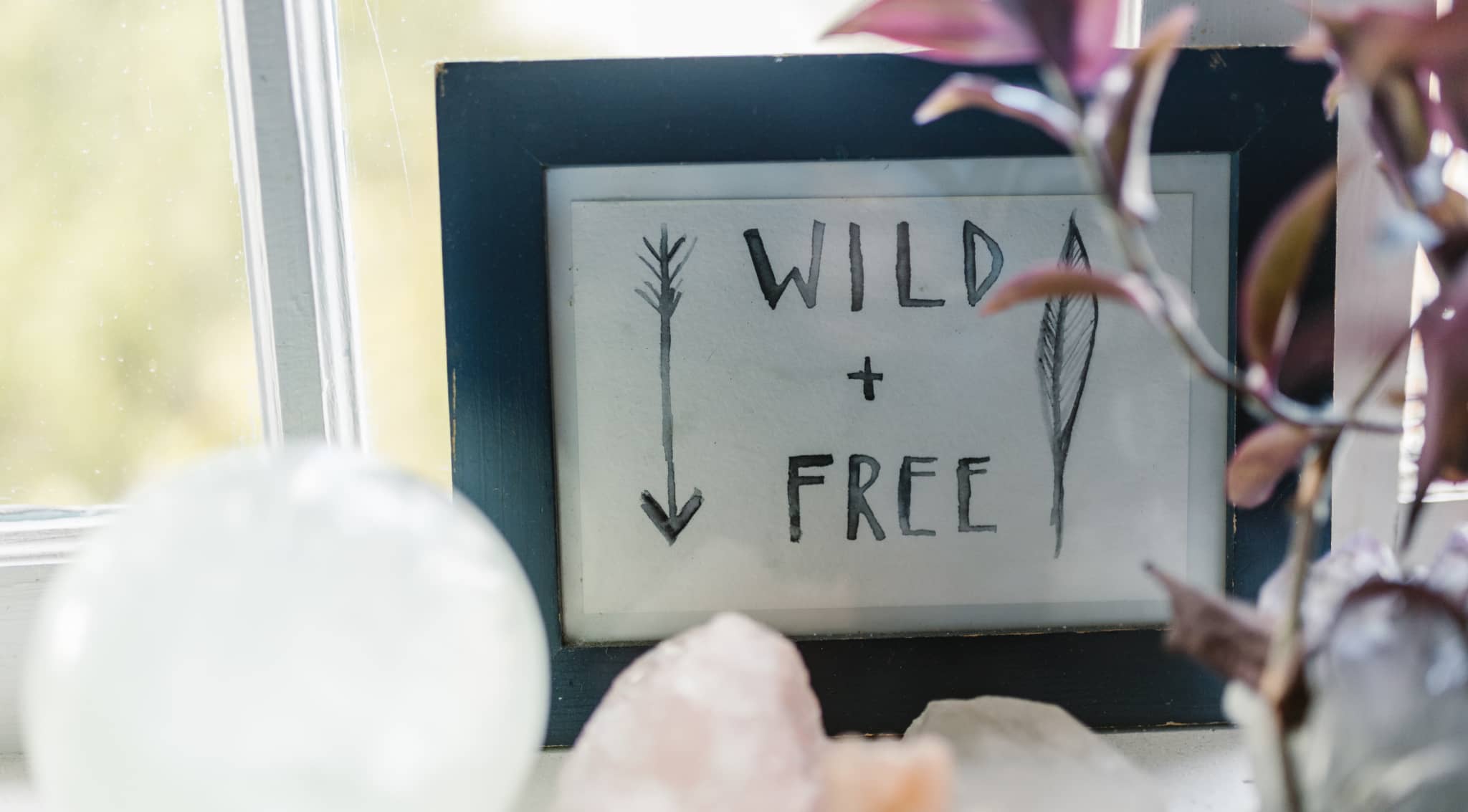 Blue picture frame with crystals and flowers around it, the frame holding the text "WILD + FREE" and an arrow on watercolour paper