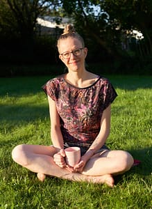 Lisa Jara sitting cross-legged on a green lawn, smiling into the camera with a lightly tilted head and a soft pink coffee mug saying "Perfectly Imperfect!"