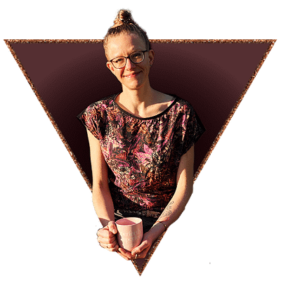 Lisa Jara smiles into the camera with a lightly tilted head, holding a soft pink coffee mug saying "Perfectly Imperfect!". Behind her is a dark red background and there's a rosegold coloured triangular frame around her.