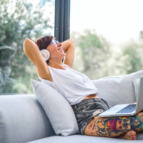 Hellhäutige Person mit großem, bunten Tattoo auf dem rechten Oberschenkel, ruht sich auf einem Sofa aus, Hände hinter dem Kopf verschränkt, Kopf zur Decke gerichtet und lächelnd