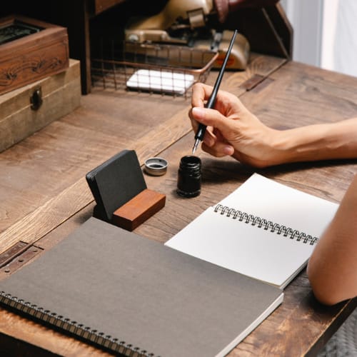 Stationary on old-fashioned desk, person's hand dipping a fountain pen into ink