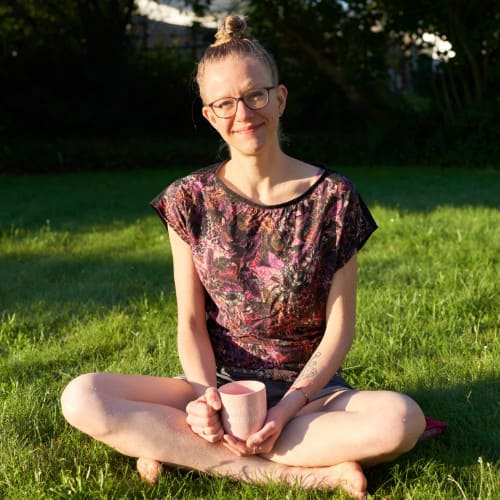 Photo of Lisa, a white woman in a dark patterned shirt, hair curled up on top of the head, sitting cross-legged on the lawn, holding a soft pink cup saying "perfectly imperfect" in her hands