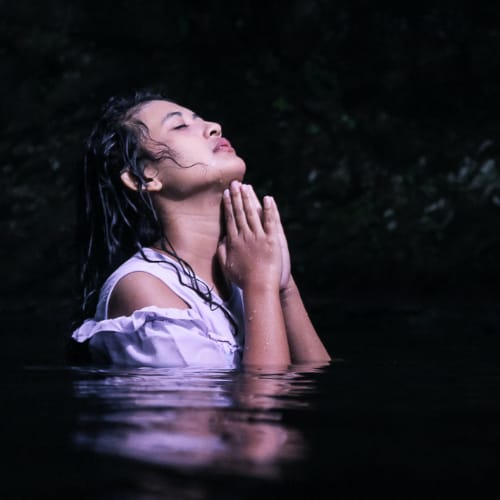 Person read as Asian female in dark waters, head lifted up, eyes closed, hands in prayer position underneath their chin